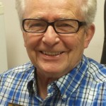 Smiling elderly man wearing glasses and a blue plaid shirt.