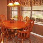 Cozy wooden dining table set by large windows with blinds.