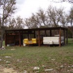 A rustic outdoor shelter with farm equipment and trailers beneath it.