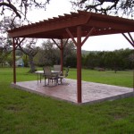 A wooden gazebo with outdoor furniture on a stone patio in a grassy yard.