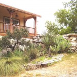 A rustic house with a wooden balcony surrounded by greenery and stone landscaping.